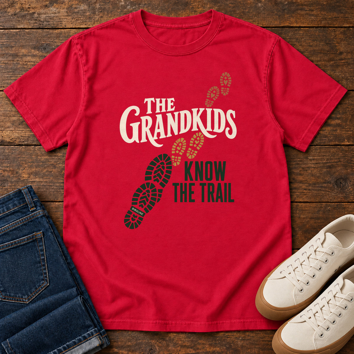 Red The Grandkids Know the Trail T-Shirt with cream and green lettering beside a trail of green, gold and brown boot prints, front flat lay on wood.