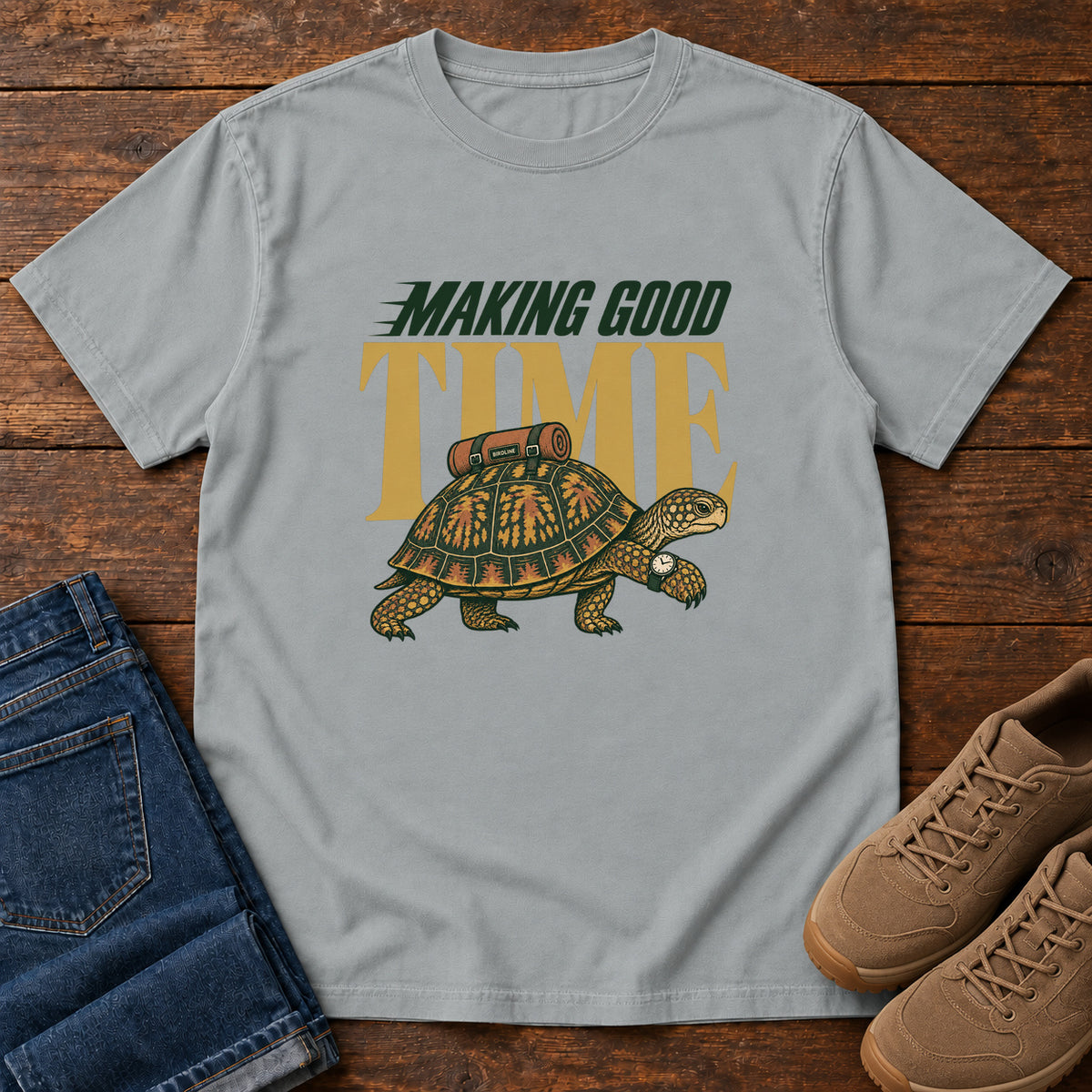 Granite Making Good Time T-Shirt with a green-and-gold turtle carrying a bedroll beneath Making Good Time lettering, front flat lay on wood.