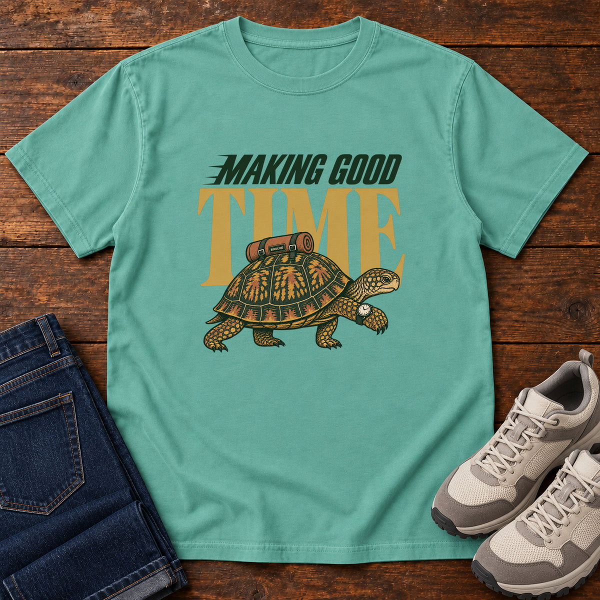 Seafoam Making Good Time T-Shirt with a green-and-gold turtle carrying a bedroll beneath Making Good Time lettering, front flat lay on wood.