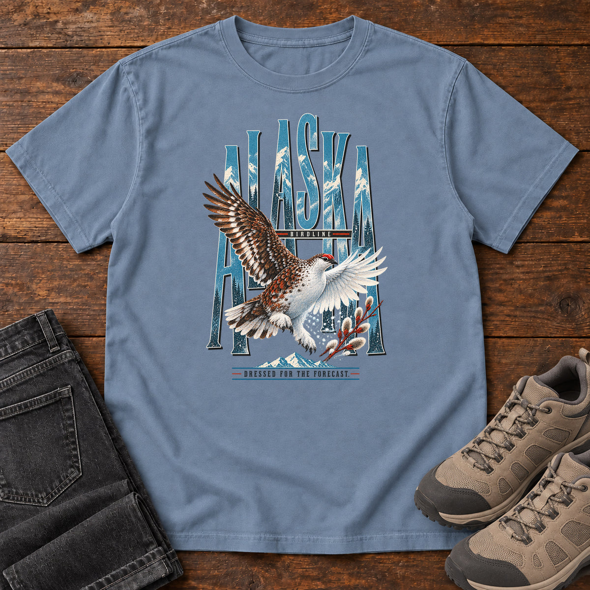 Blue Jean Alaska Dressed for the Forecast Eagle T-Shirt with a white-and-brown bird with raised wings over Alaska lettering and snowy mountains; front flat lay on wood.