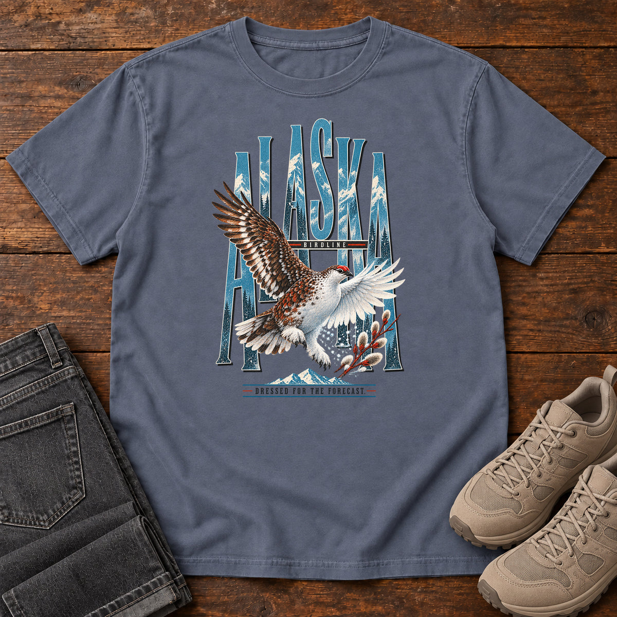 Denim Alaska Dressed for the Forecast Eagle T-Shirt with a white-and-brown bird with raised wings over Alaska lettering and snowy mountains; front flat lay on wood.