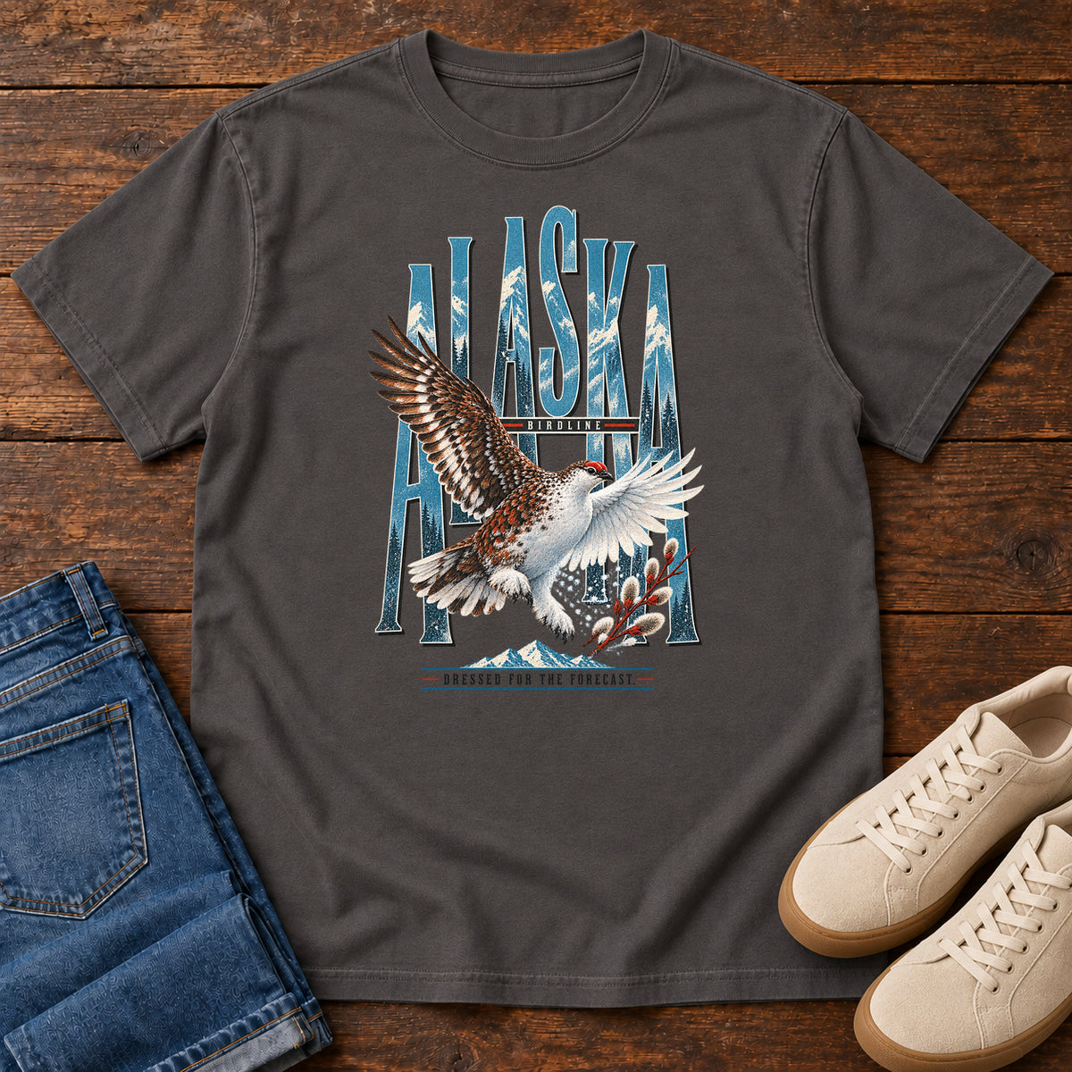 Graphite Alaska Dressed for the Forecast Eagle T-Shirt with a white-and-brown bird with raised wings over Alaska lettering and snowy mountains; front flat lay on wood.