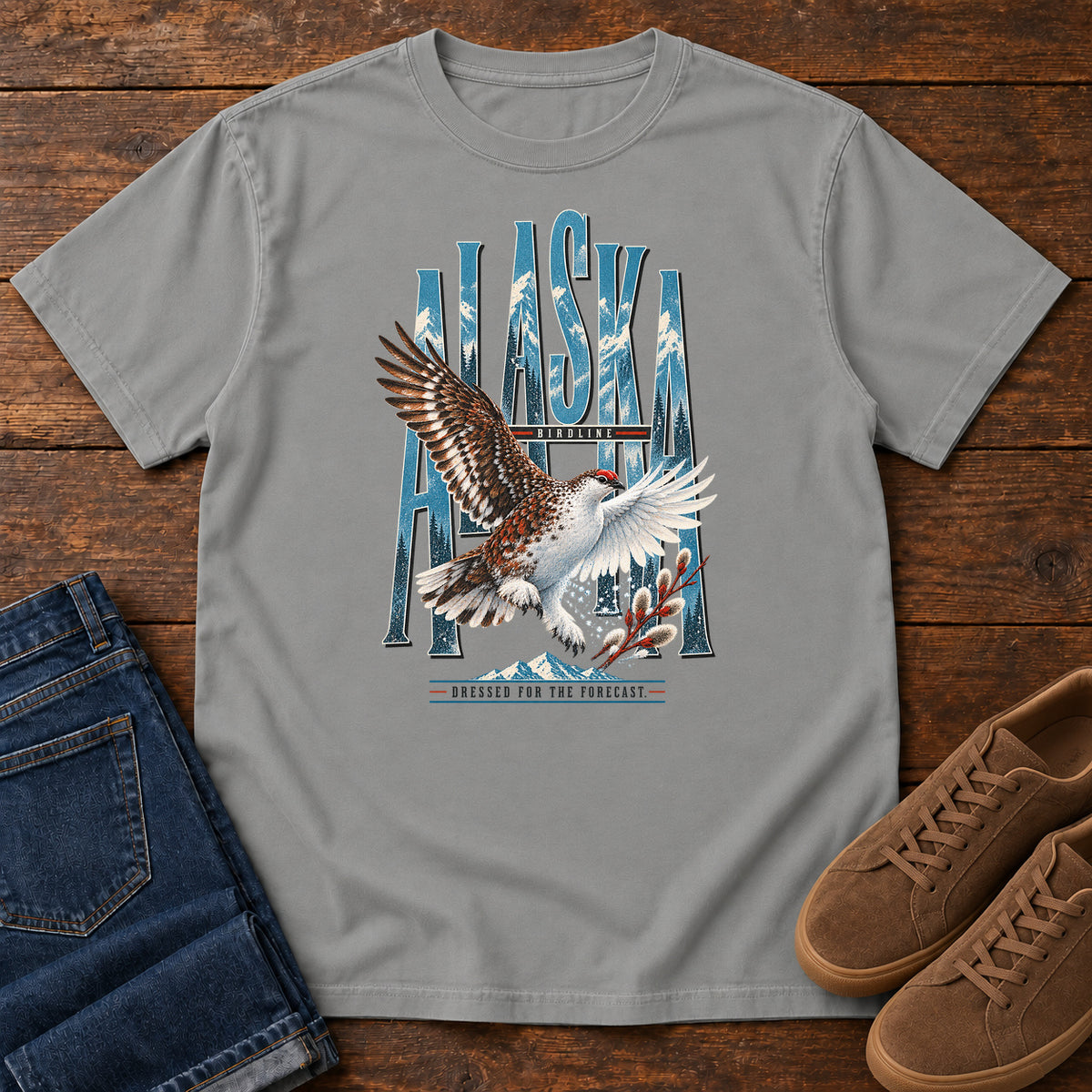 Grey Alaska Dressed for the Forecast Eagle T-Shirt with a white-and-brown bird with raised wings over Alaska lettering and snowy mountains; front flat lay on wood.