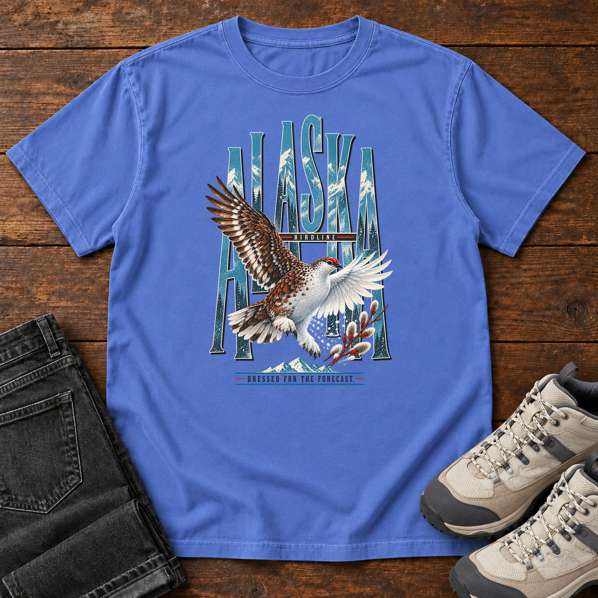 Mystic Blue Alaska Dressed for the Forecast Eagle T-Shirt with a white-and-brown bird with raised wings over Alaska lettering and snowy mountains; front flat lay on wood.