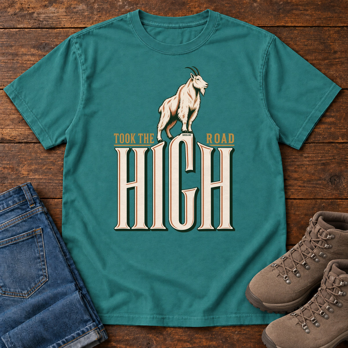 Took the High Road T-Shirt in Emerald, front laid flat on wood, featuring a white mountain goat standing above oversized cream HIGH lettering.