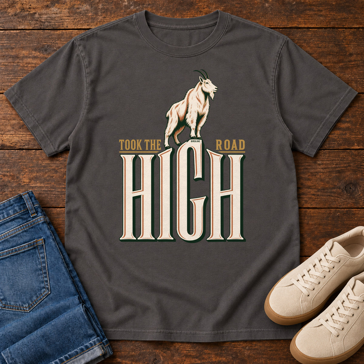 Took the High Road T-Shirt in Graphite, front laid flat on wood, featuring a white mountain goat standing above oversized cream HIGH lettering.