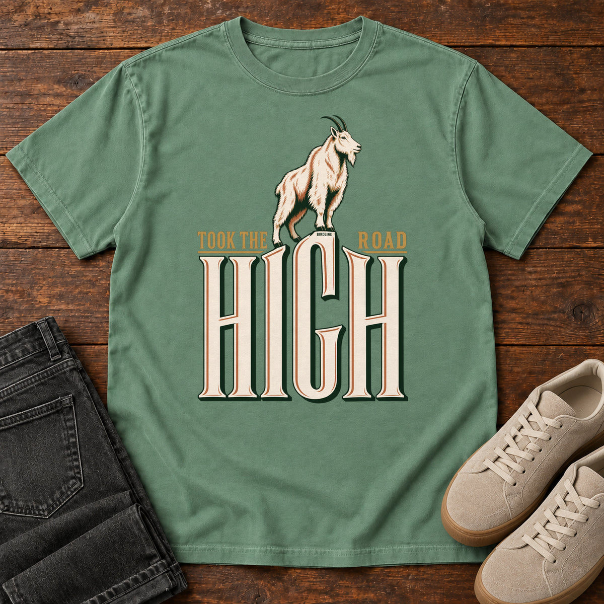 Took the High Road T-Shirt in Light Green, front laid flat on wood, featuring a white mountain goat standing above oversized cream HIGH lettering.