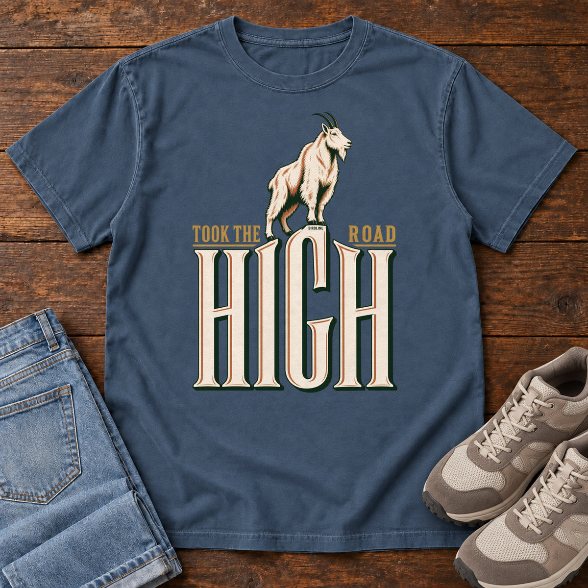 Took the High Road T-Shirt in Midnight, front laid flat on wood, featuring a white mountain goat standing above oversized cream HIGH lettering.
