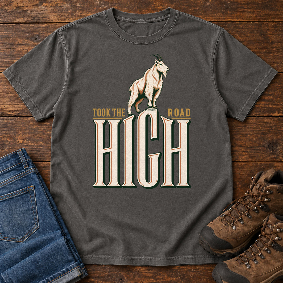 Took the High Road T-Shirt in Pepper, front laid flat on wood, featuring a white mountain goat standing above oversized cream HIGH lettering.