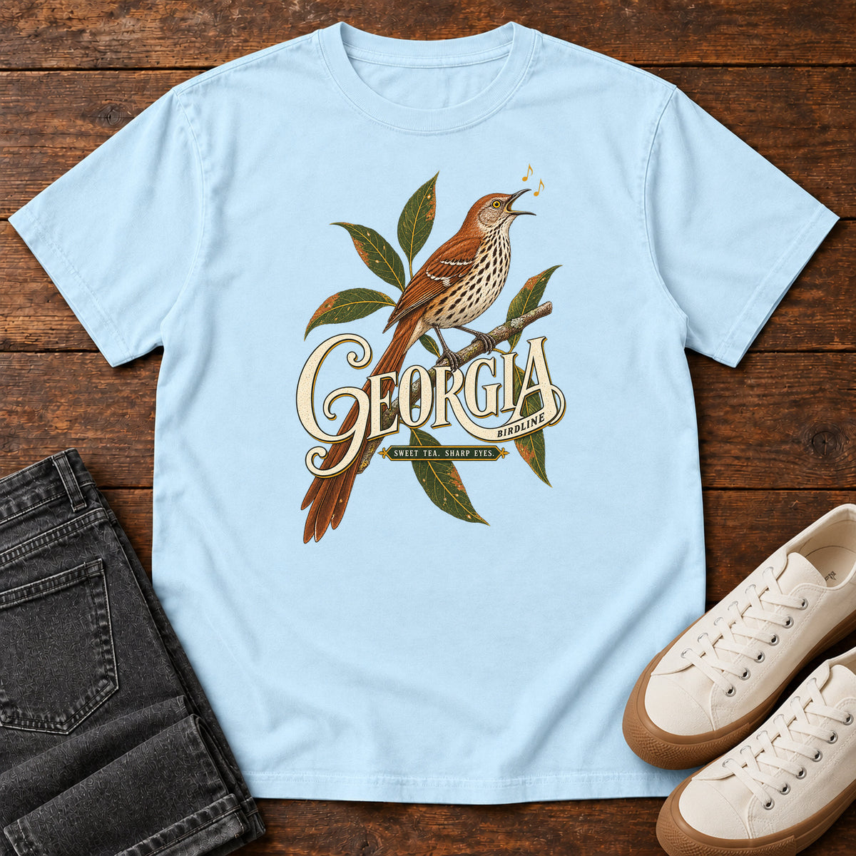 Chambray Georgia Sharp Eyes T-Shirt with a brown, speckle-breasted bird on a leafy branch above Georgia lettering, front flat lay on wood.