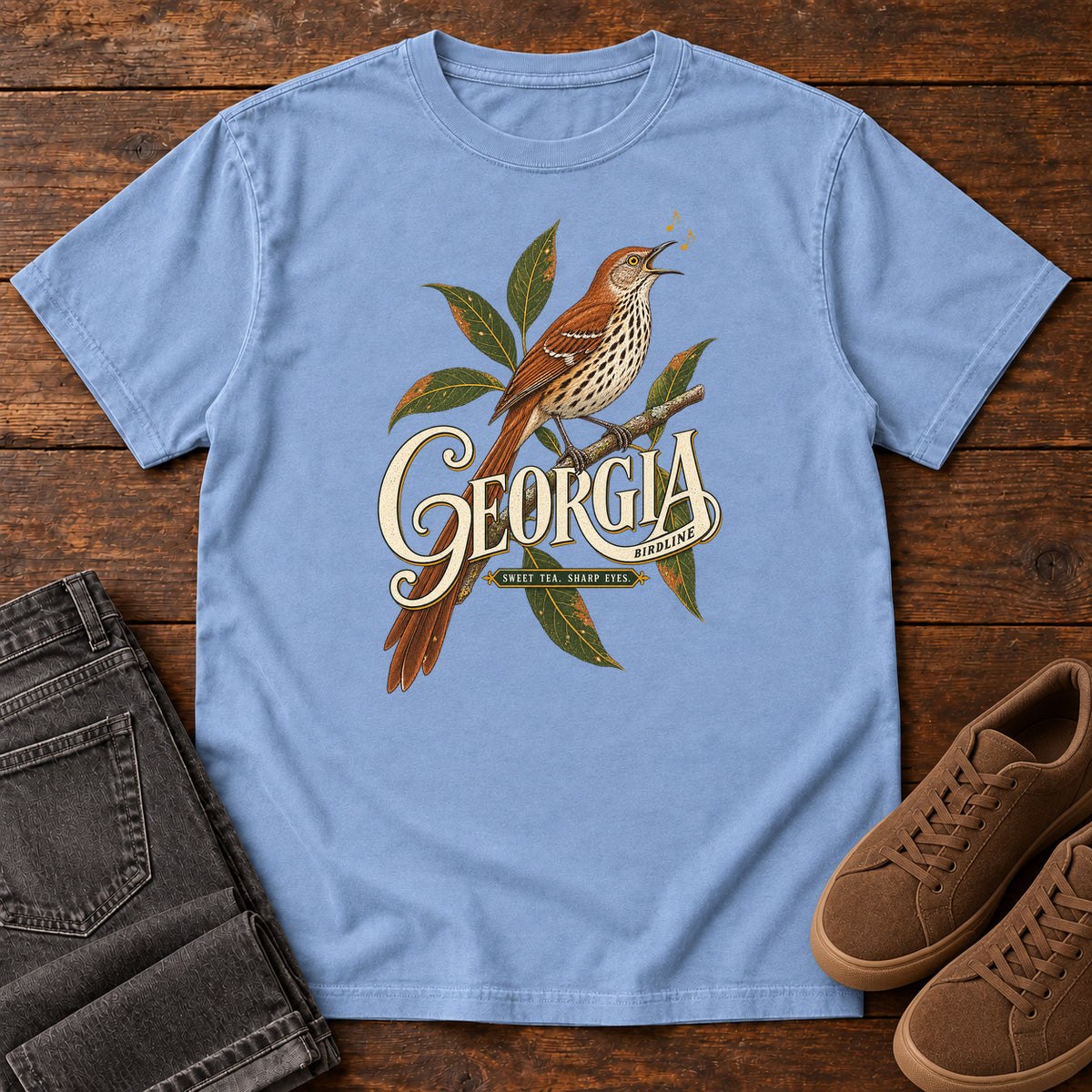 Washed Denim Georgia Sharp Eyes T-Shirt with a brown, speckle-breasted bird on a leafy branch above Georgia lettering, front flat lay on wood.