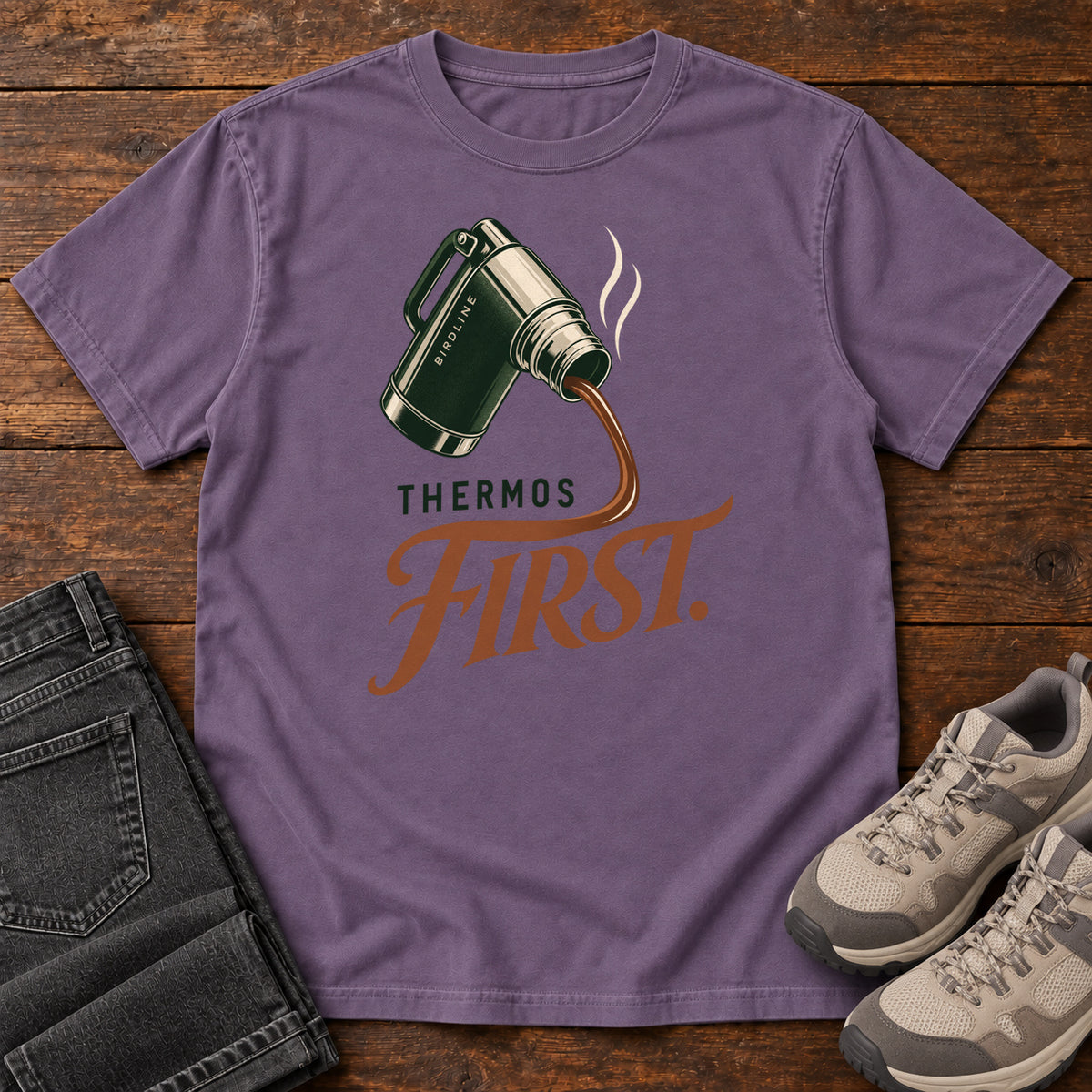 Grape Thermos First T-Shirt with a green thermos pouring a steaming drink into rust-colored script, front flat lay on wood.