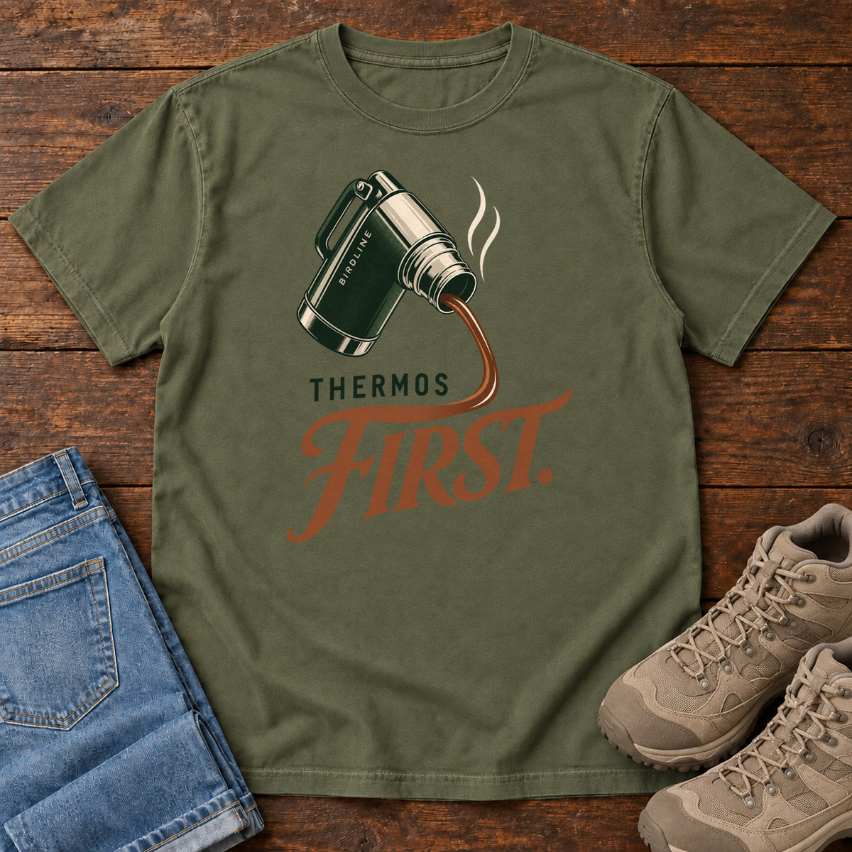 Hemp Thermos First T-Shirt with a green thermos pouring a steaming drink into rust-colored script, front flat lay on wood.