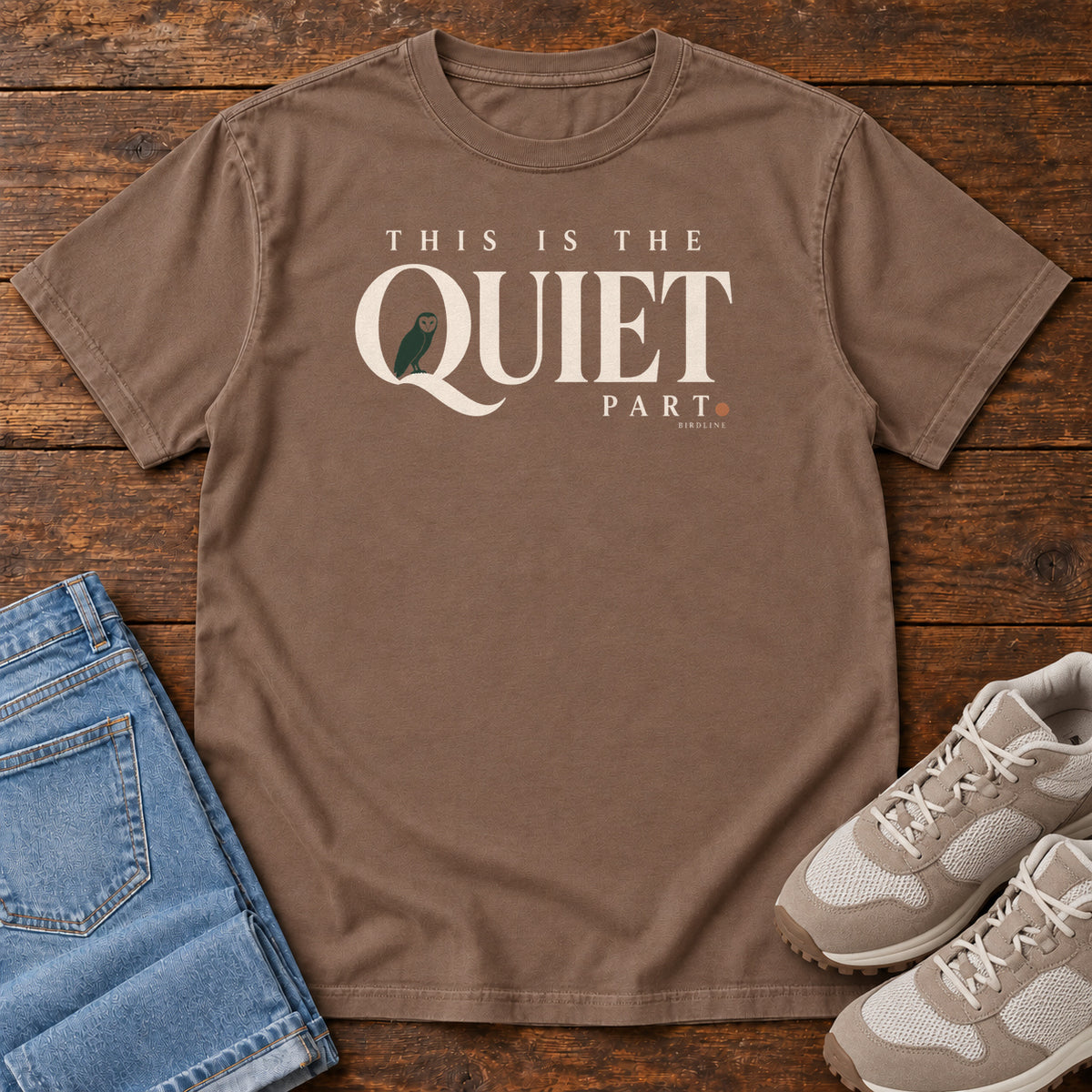 Espresso This Is the Quiet Part T-Shirt with cream This Is the Quiet Part lettering with a small green bird inside the Q, front flat lay on wood.
