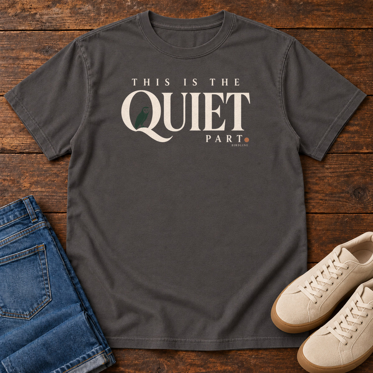 Graphite This Is the Quiet Part T-Shirt with cream This Is the Quiet Part lettering with a small green bird inside the Q, front flat lay on wood.