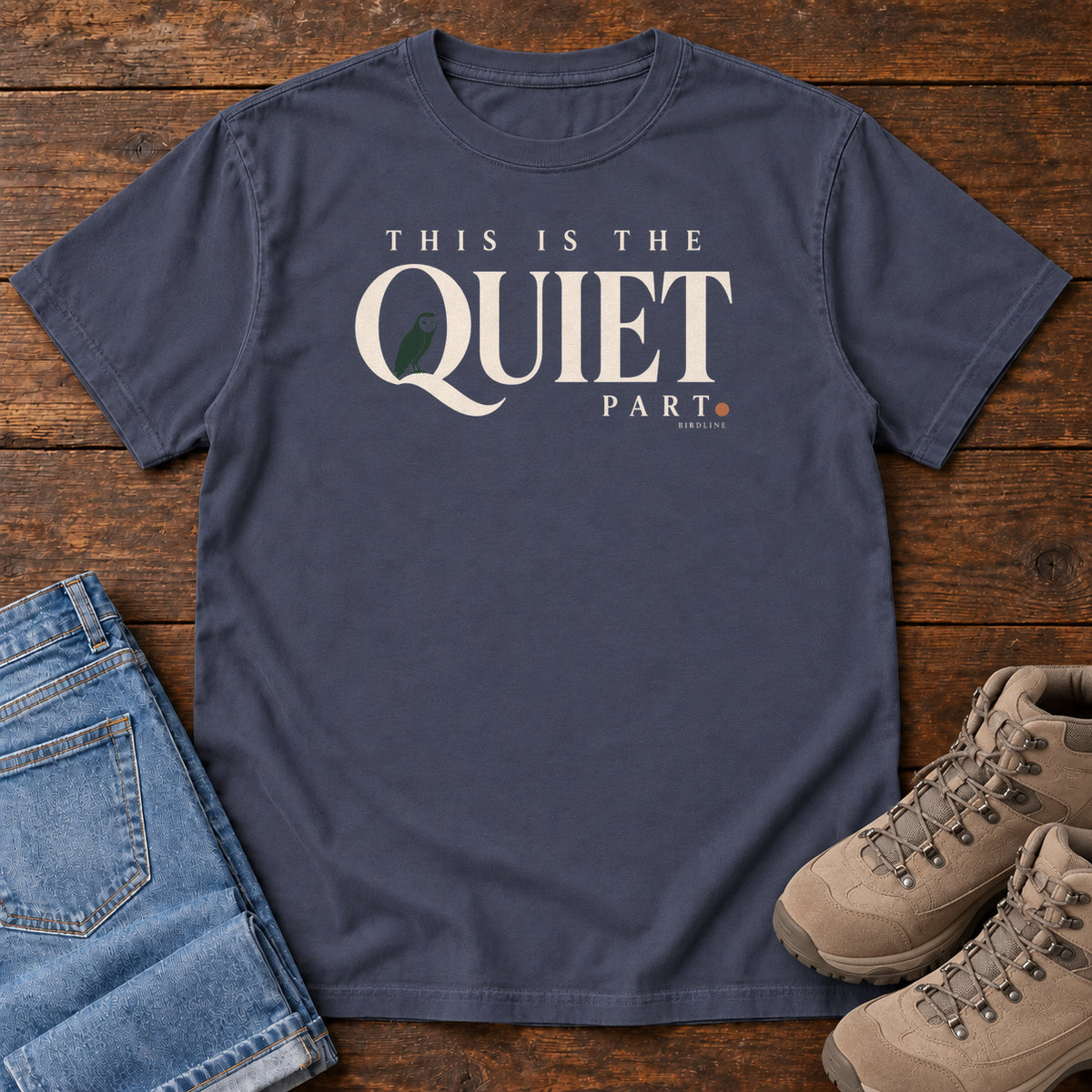 Navy This Is the Quiet Part T-Shirt with cream This Is the Quiet Part lettering with a small green bird inside the Q, front flat lay on wood.
