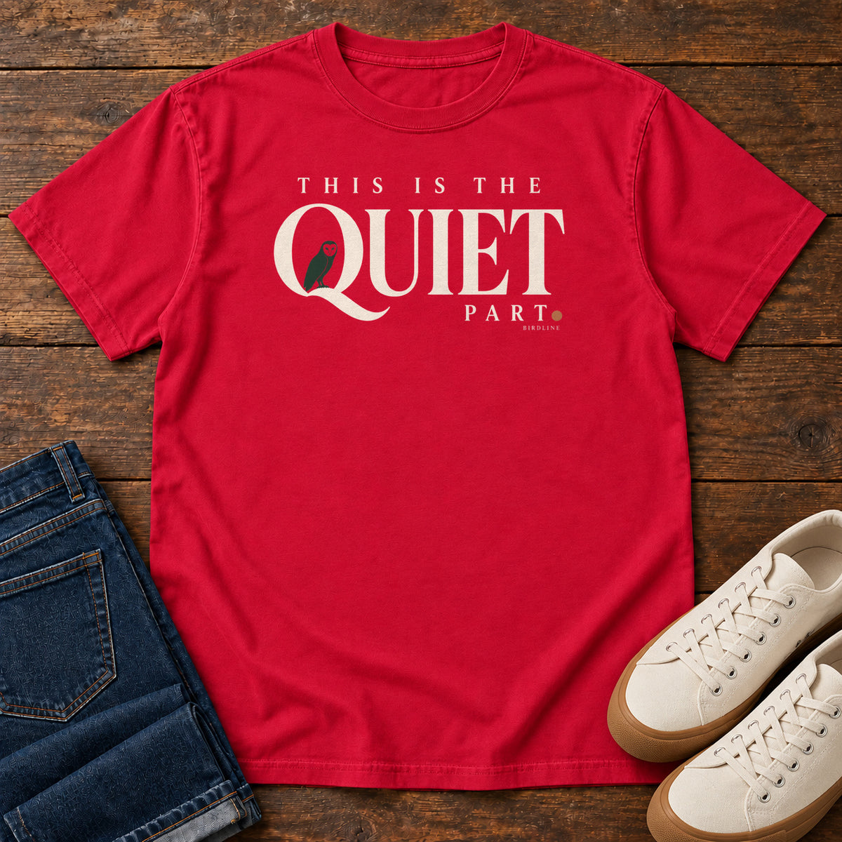 Red This Is the Quiet Part T-Shirt with cream This Is the Quiet Part lettering with a small green bird inside the Q, front flat lay on wood.