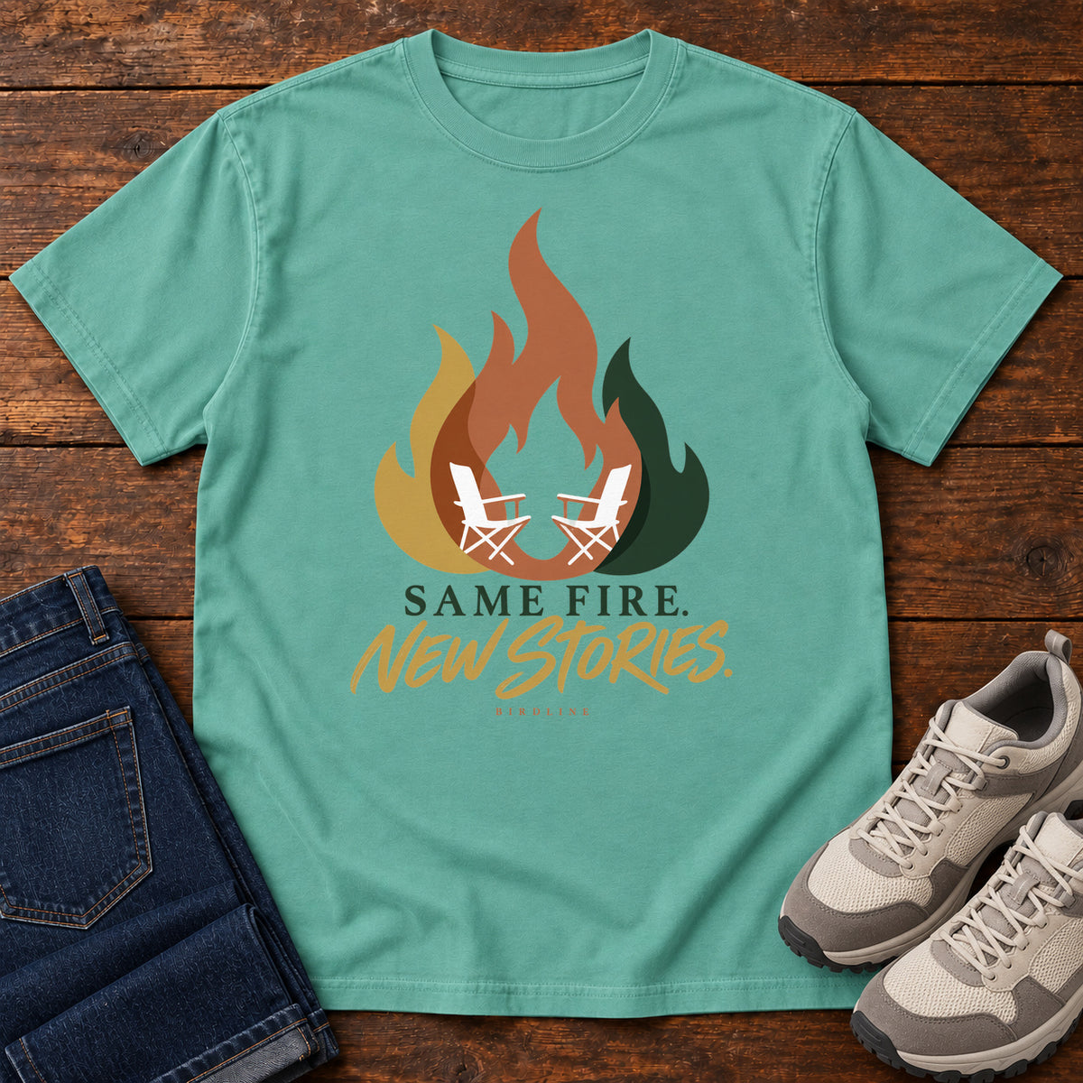 Seafoam Same Fire New Stories T-Shirt with a stylized orange, gold and green flame framing two white camp chairs; front flat lay on wood.