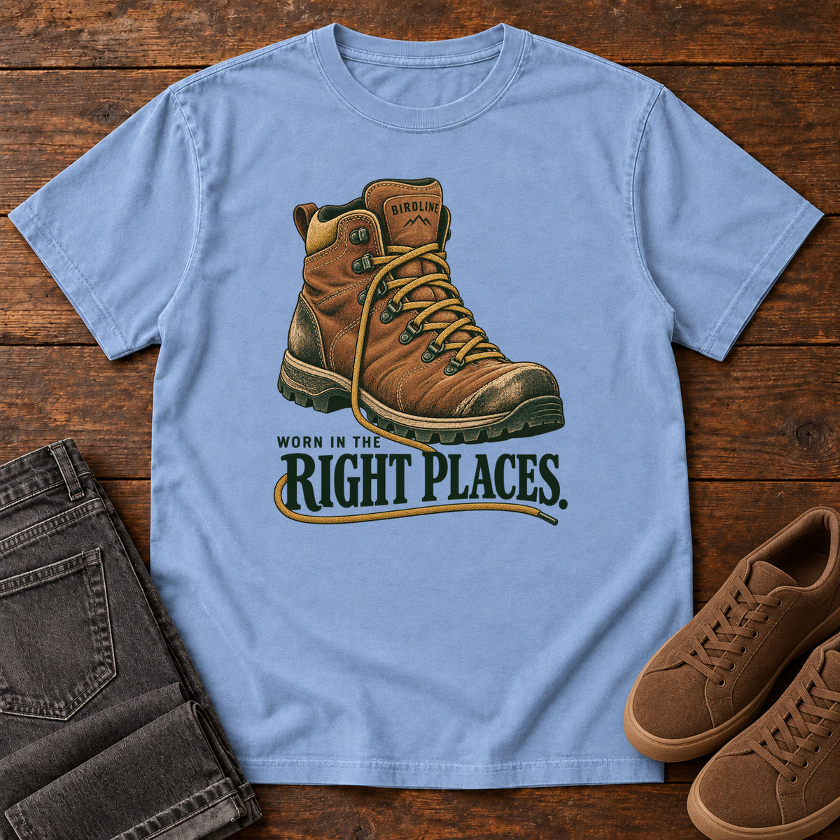Worn in the Right Places T-Shirt in Washed Denim, front laid flat on wood, featuring a brown lace-up hiking boot above dark green slogan lettering.