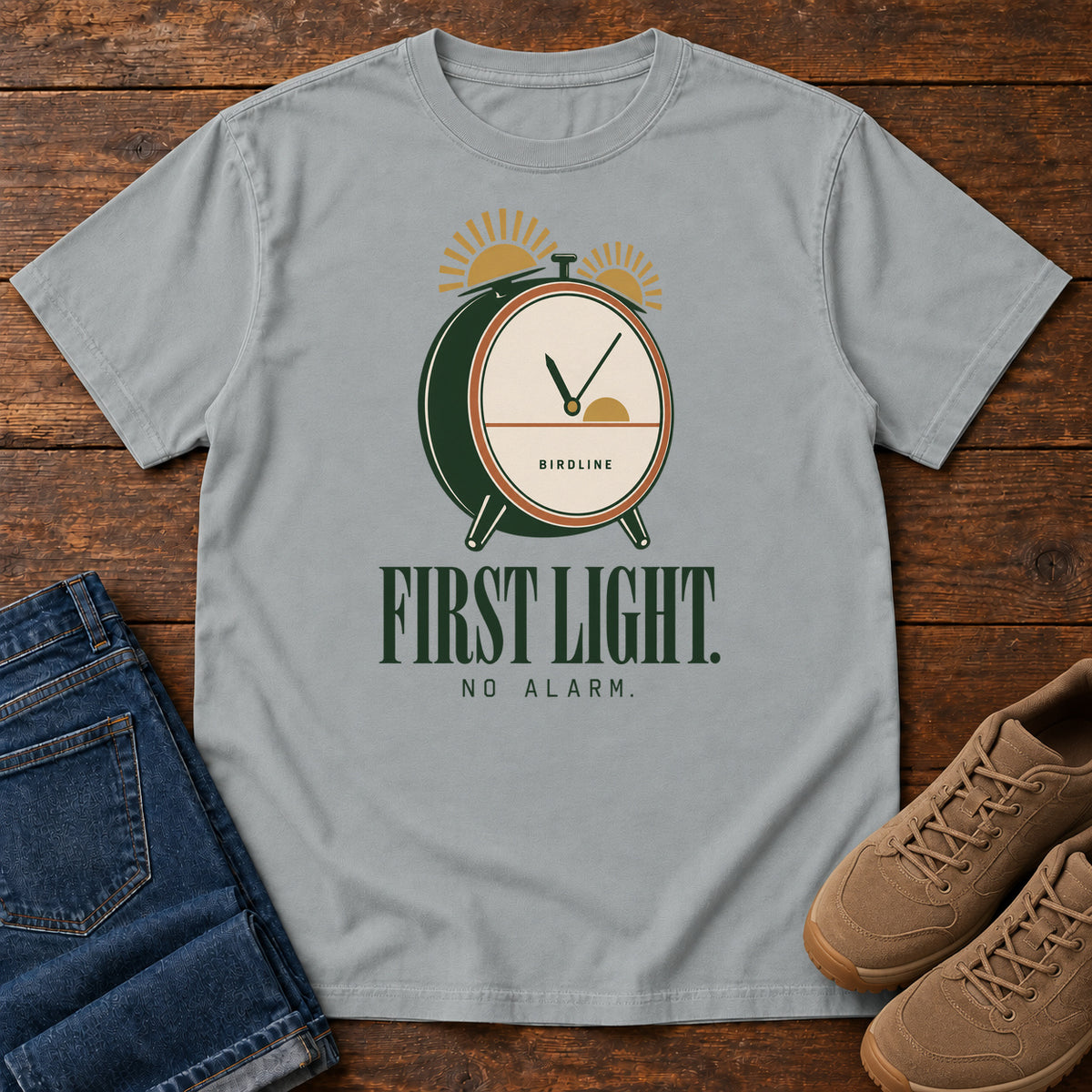 Granite First Light No Alarm T-Shirt with a green-and-cream clock with gold sunrise shapes above First Light No Alarm lettering, front flat lay on wood.