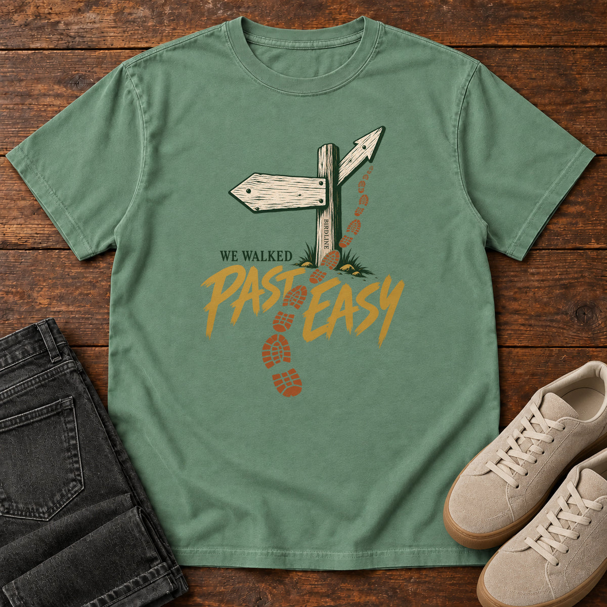 Light Green We Walked Past Easy T-Shirt with a wooden trail sign, orange boot prints and golden script, front flat lay on wood.