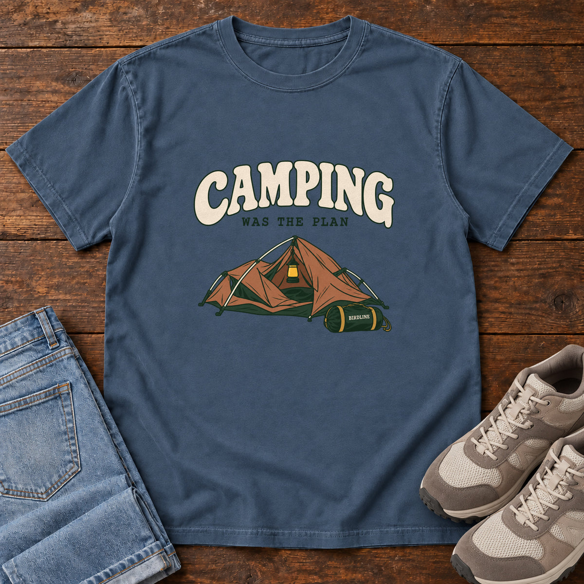 Midnight Camping Was the Plan T-Shirt with an orange tent with a hanging lantern and green gear bag; front flat lay on wood.