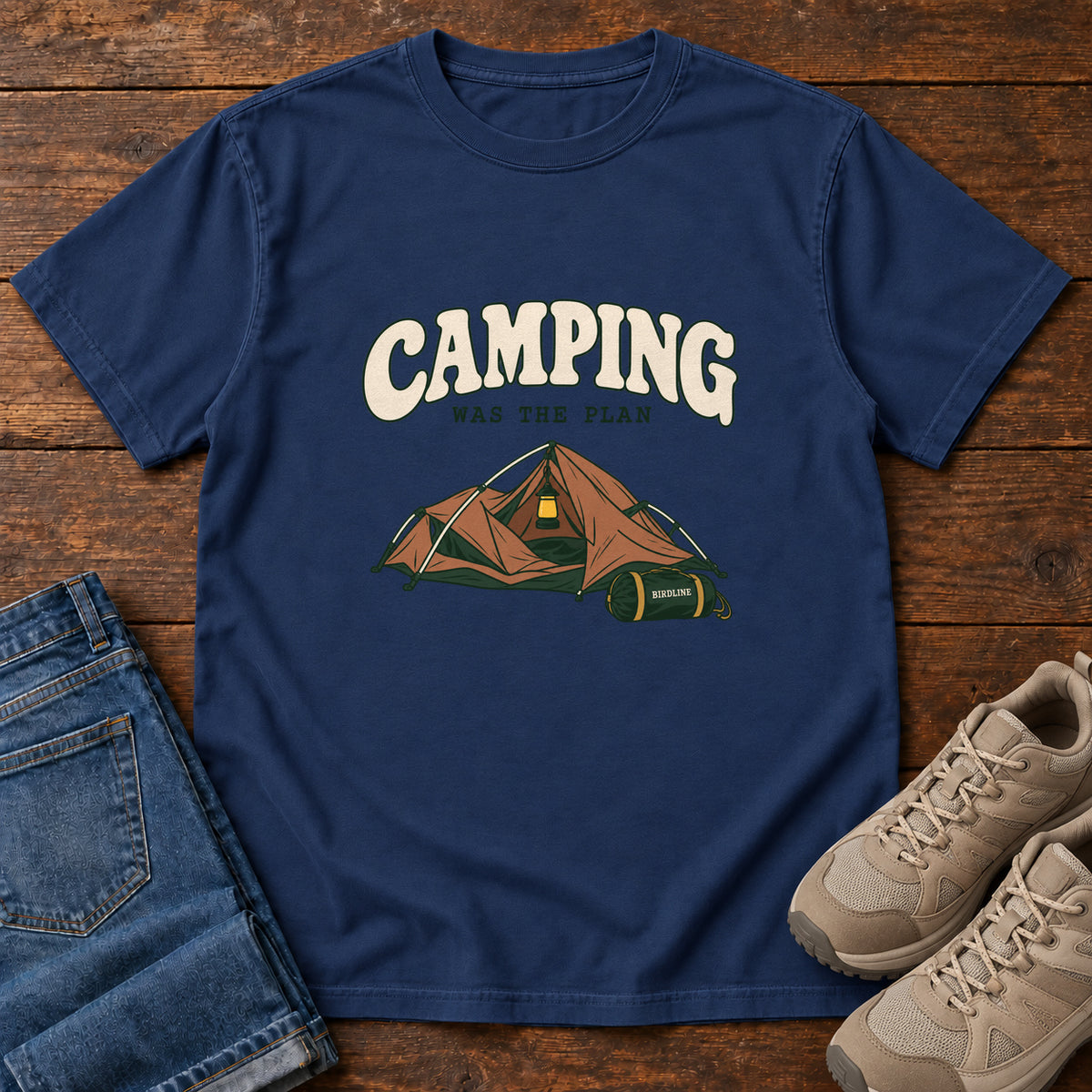 True Navy Camping Was the Plan T-Shirt with an orange tent with a hanging lantern and green gear bag; front flat lay on wood.