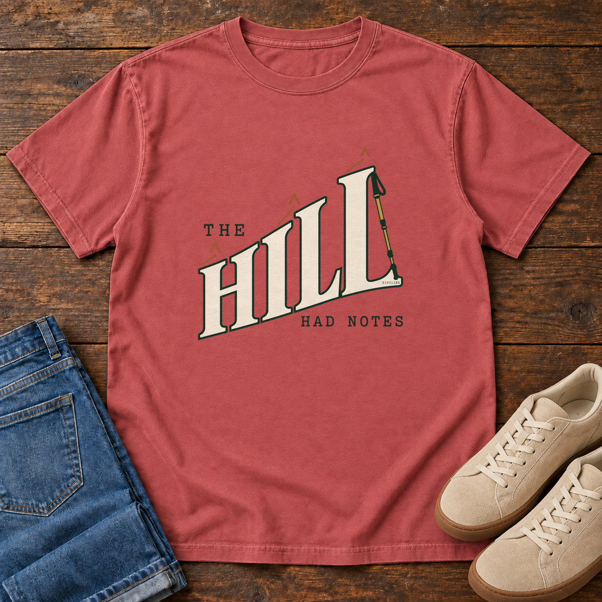The Hill Had Notes T-Shirt in Crimson, front laid flat on wood, featuring ascending cream HILL lettering with small orange chevrons and a trekking pole.