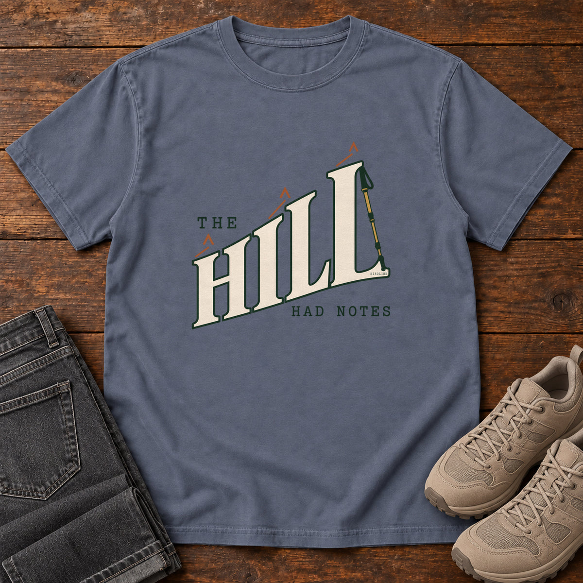 The Hill Had Notes T-Shirt in Denim, front laid flat on wood, featuring ascending cream HILL lettering with small orange chevrons and a trekking pole.