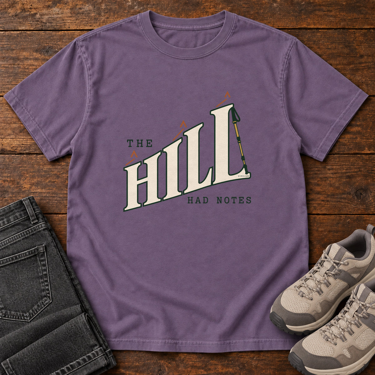 The Hill Had Notes T-Shirt in Grape, front laid flat on wood, featuring ascending cream HILL lettering with small orange chevrons and a trekking pole.