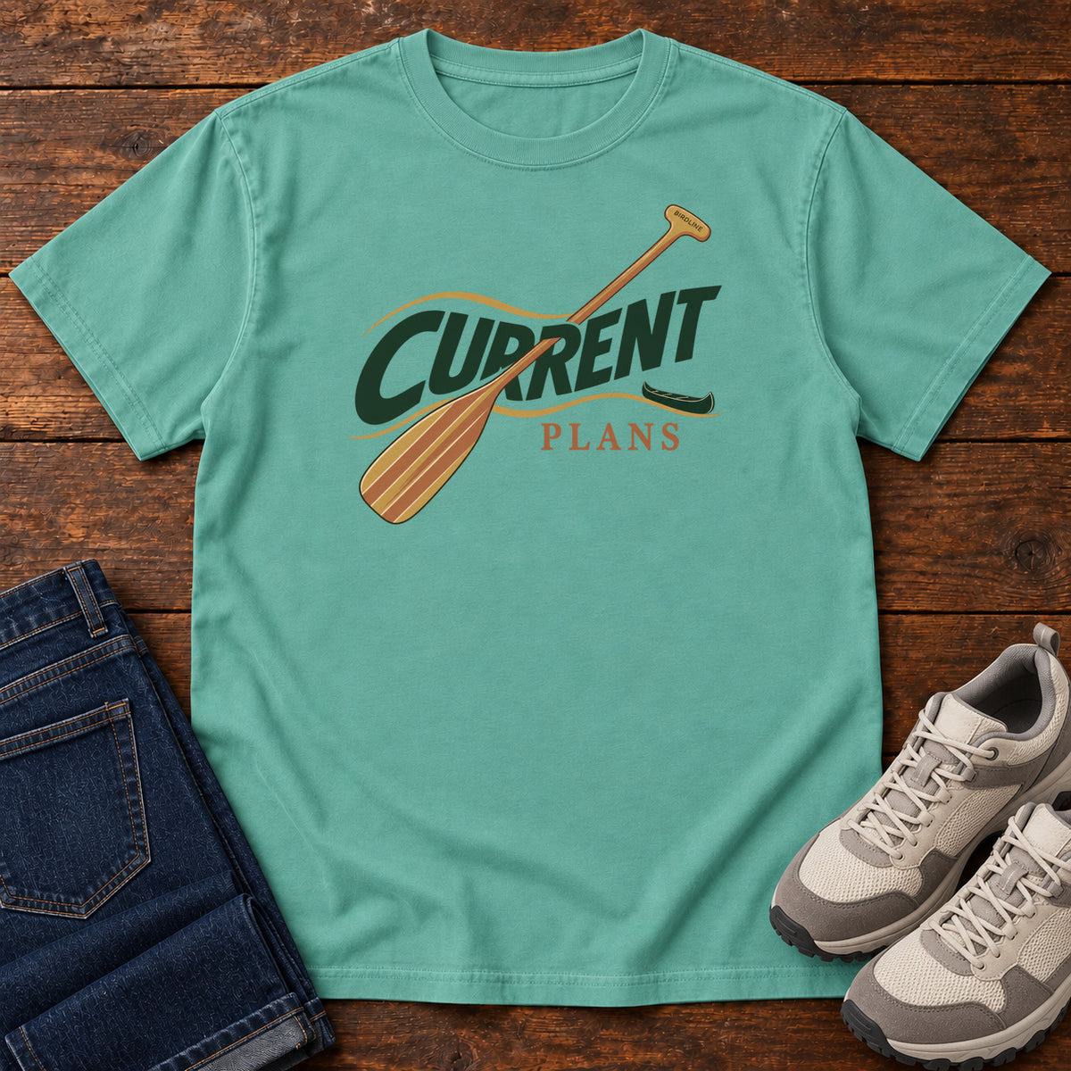 Seafoam Current Plans T-Shirt with a diagonal wooden paddle crossing green and gold Current Plans lettering; front flat lay on wood.