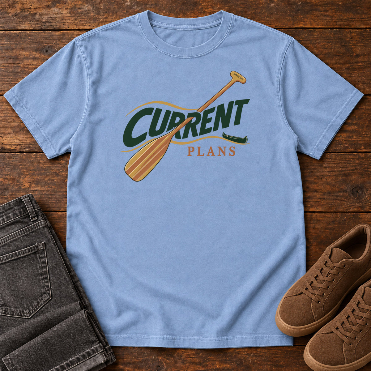 Washed Denim Current Plans T-Shirt with a diagonal wooden paddle crossing green and gold Current Plans lettering; front flat lay on wood.