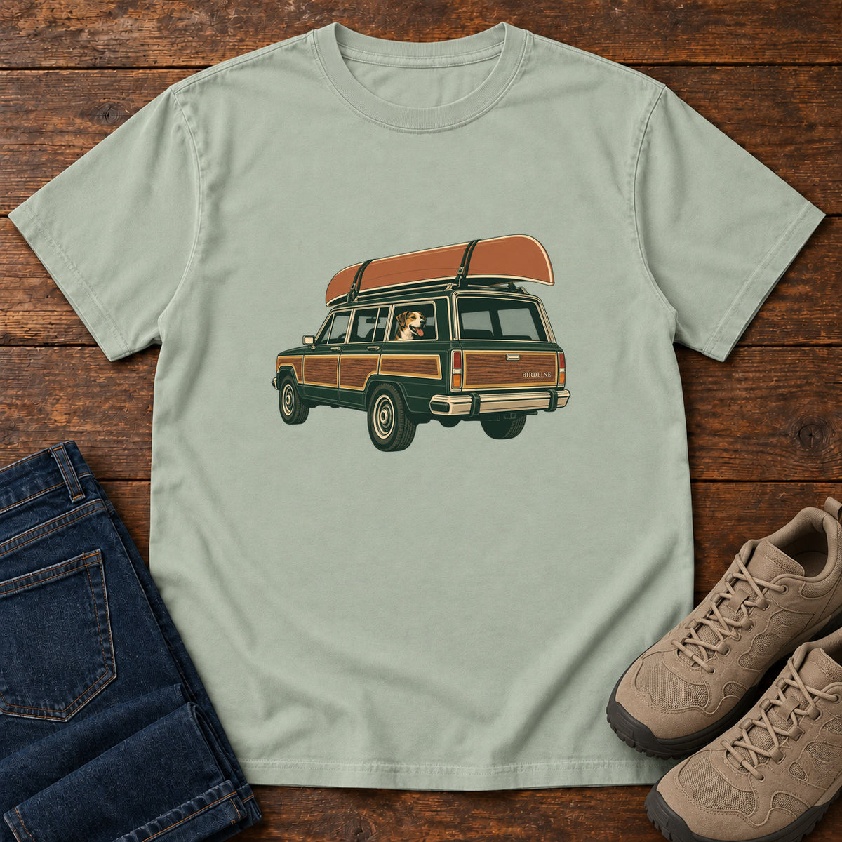 Bay Canoe Wagon and Dog T-Shirt with a green wood-paneled wagon, roof-mounted canoe and dog in the window, front flat lay on wood.