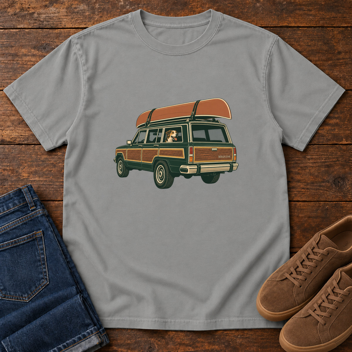 Grey Canoe Wagon and Dog T-Shirt with a green wood-paneled wagon, roof-mounted canoe and dog in the window, front flat lay on wood.