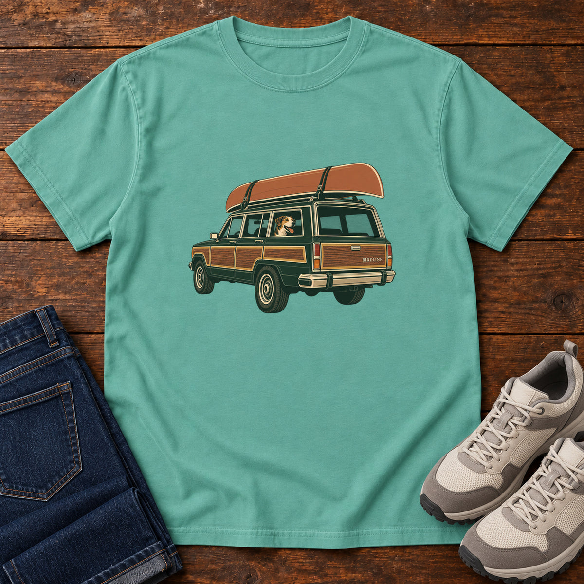 Seafoam Canoe Wagon and Dog T-Shirt with a green wood-paneled wagon, roof-mounted canoe and dog in the window, front flat lay on wood.