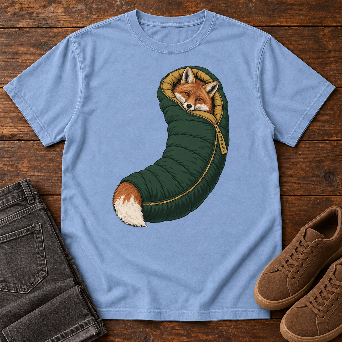 Washed Denim Fox in a Sleeping Bag T-Shirt with an orange fox tucked into a green sleeping bag with its white-tipped tail showing; front flat lay on wood.