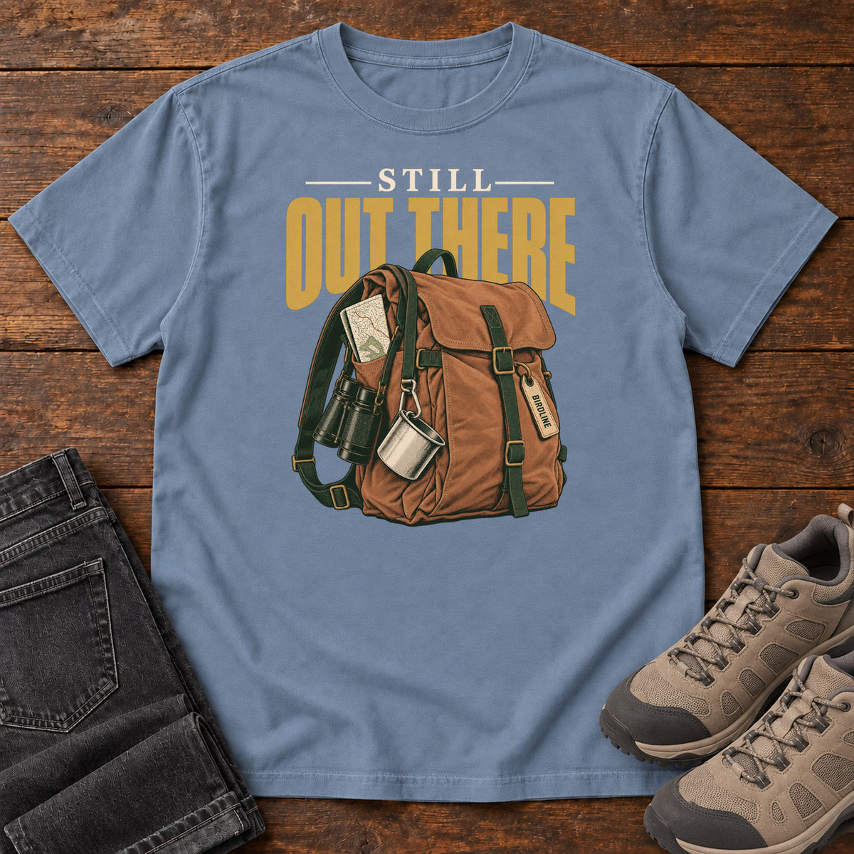 Blue Jean Still Out There T-Shirt with a brown hiking backpack with binoculars, a mug and gold lettering, front flat lay on wood.