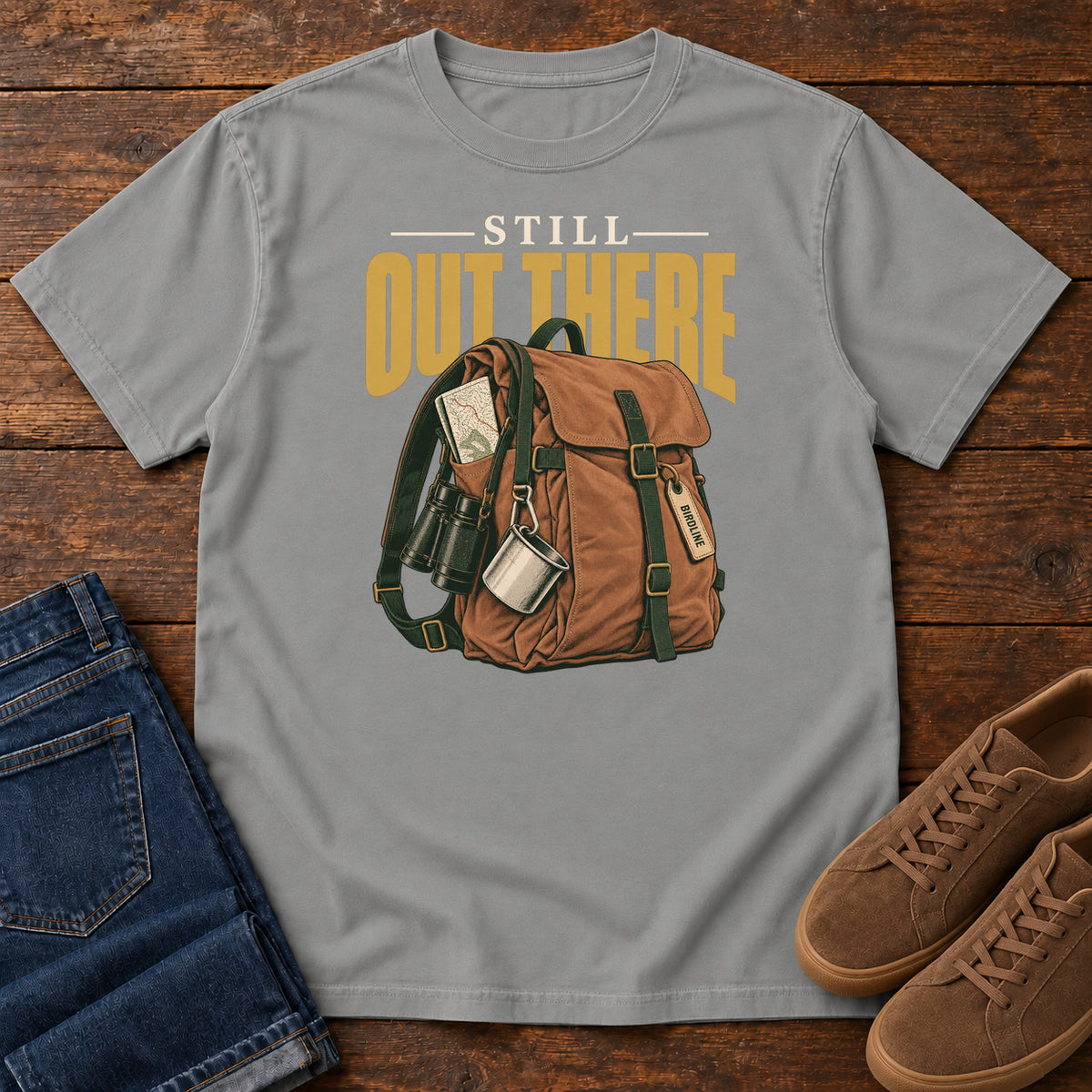 Grey Still Out There T-Shirt with a brown hiking backpack with binoculars, a mug and gold lettering, front flat lay on wood.