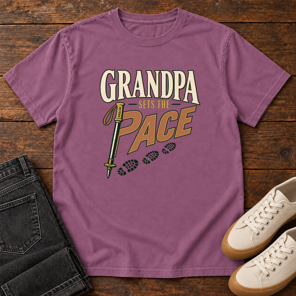 Grandpa Sets the Pace T-Shirt in Berry, front laid flat on wood, featuring cream-and-orange slogan lettering beside a trekking pole and a trail of boot prints.