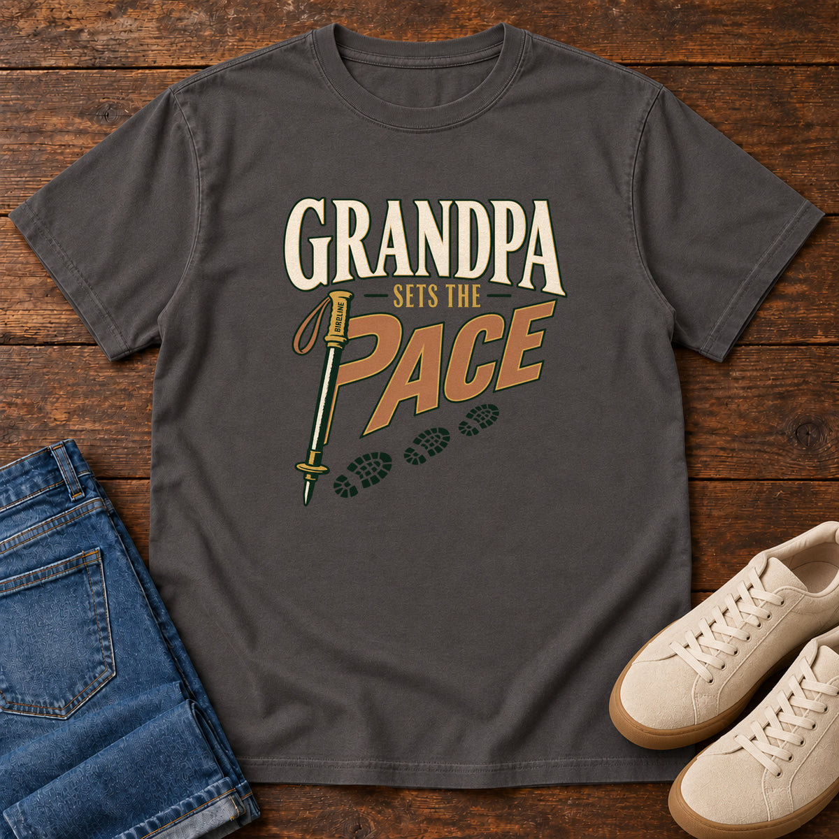 Grandpa Sets the Pace T-Shirt in Graphite, front laid flat on wood, featuring cream-and-orange slogan lettering beside a trekking pole and a trail of boot prints.