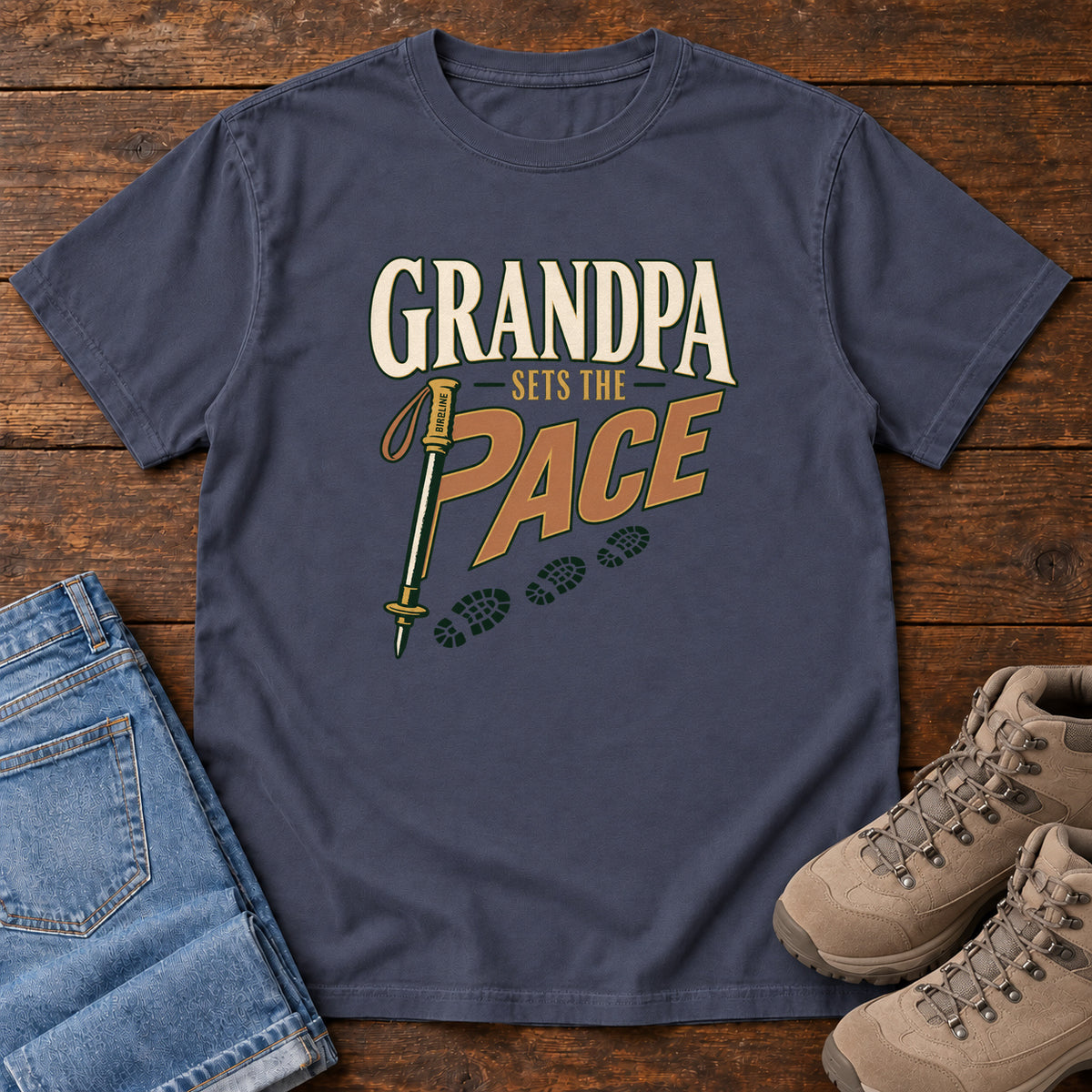 Grandpa Sets the Pace T-Shirt in Navy, front laid flat on wood, featuring cream-and-orange slogan lettering beside a trekking pole and a trail of boot prints.