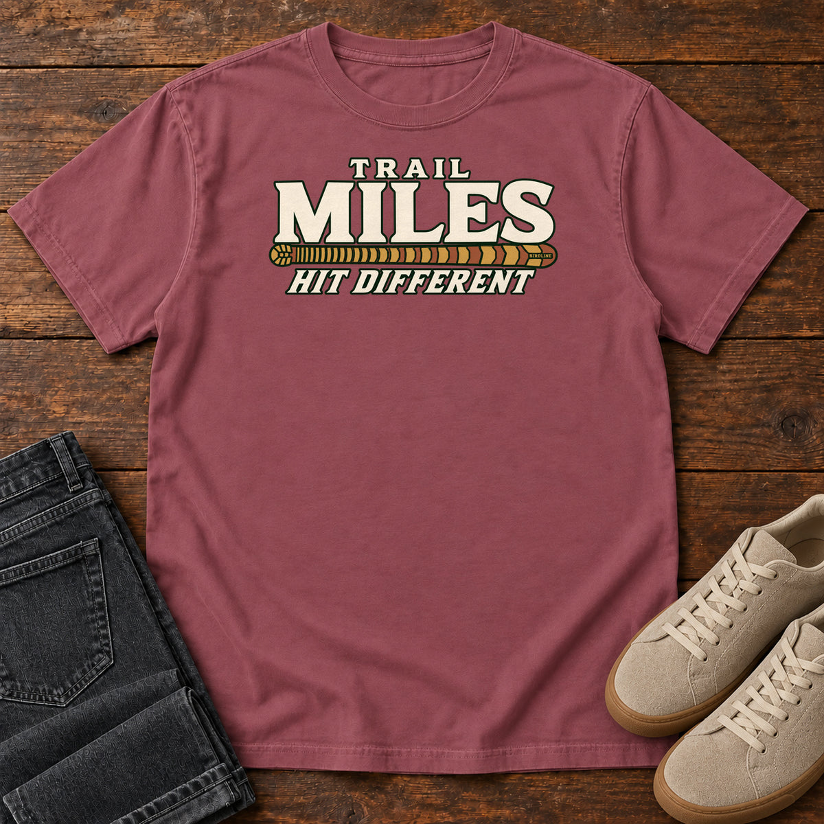 Brick Trail Miles Hit Different T-Shirt with cream lettering and a gold-to-rust patterned stripe, front flat lay on wood.