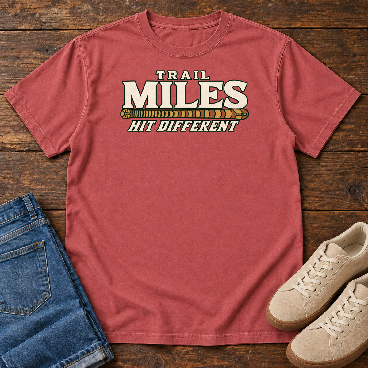 Crimson Trail Miles Hit Different T-Shirt with cream lettering and a gold-to-rust patterned stripe, front flat lay on wood.