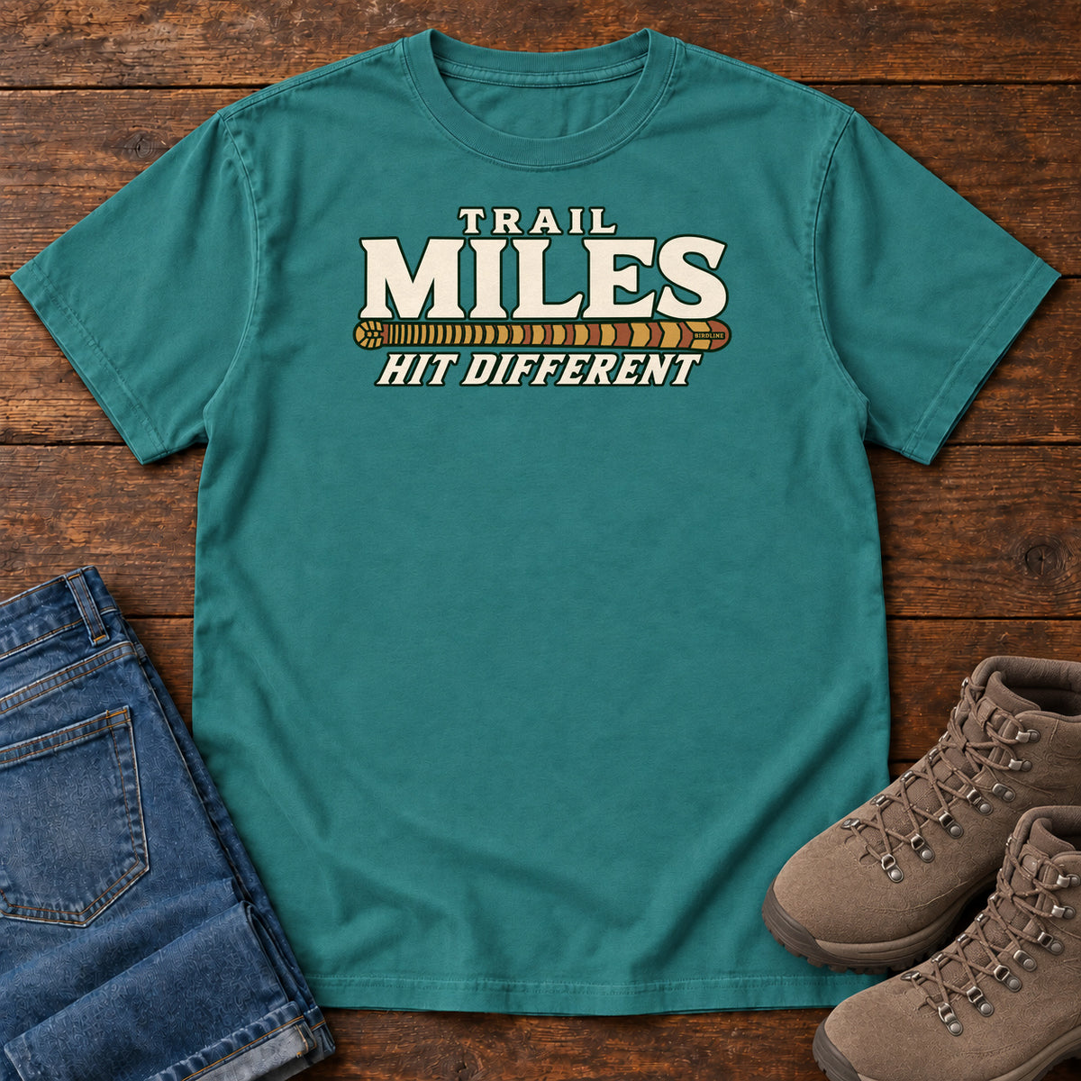 Emerald Trail Miles Hit Different T-Shirt with cream lettering and a gold-to-rust patterned stripe, front flat lay on wood.