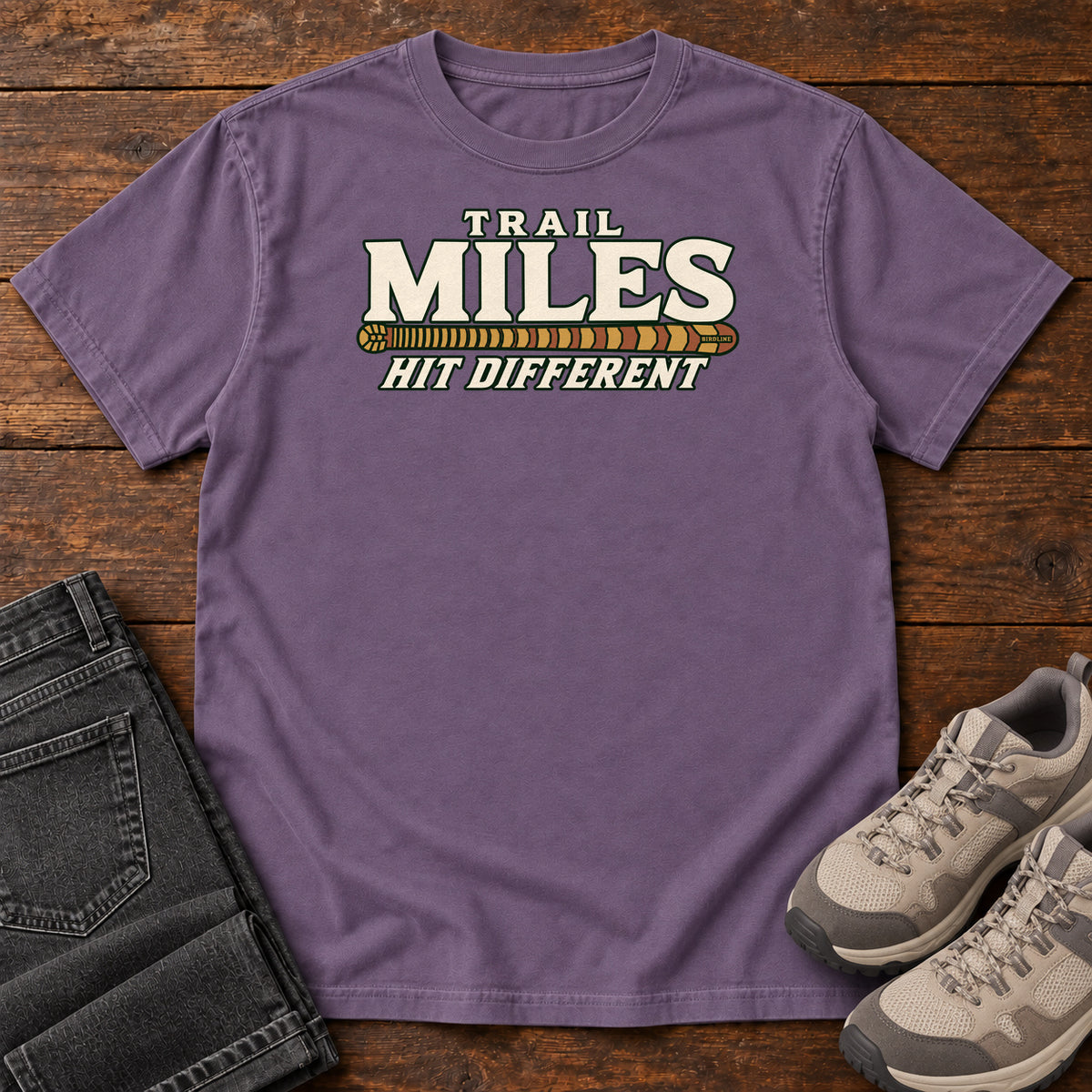 Grape Trail Miles Hit Different T-Shirt with cream lettering and a gold-to-rust patterned stripe, front flat lay on wood.