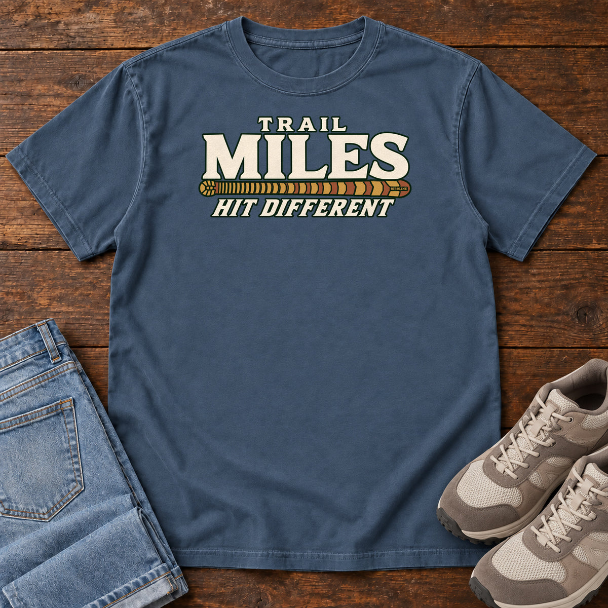 Midnight Trail Miles Hit Different T-Shirt with cream lettering and a gold-to-rust patterned stripe, front flat lay on wood.