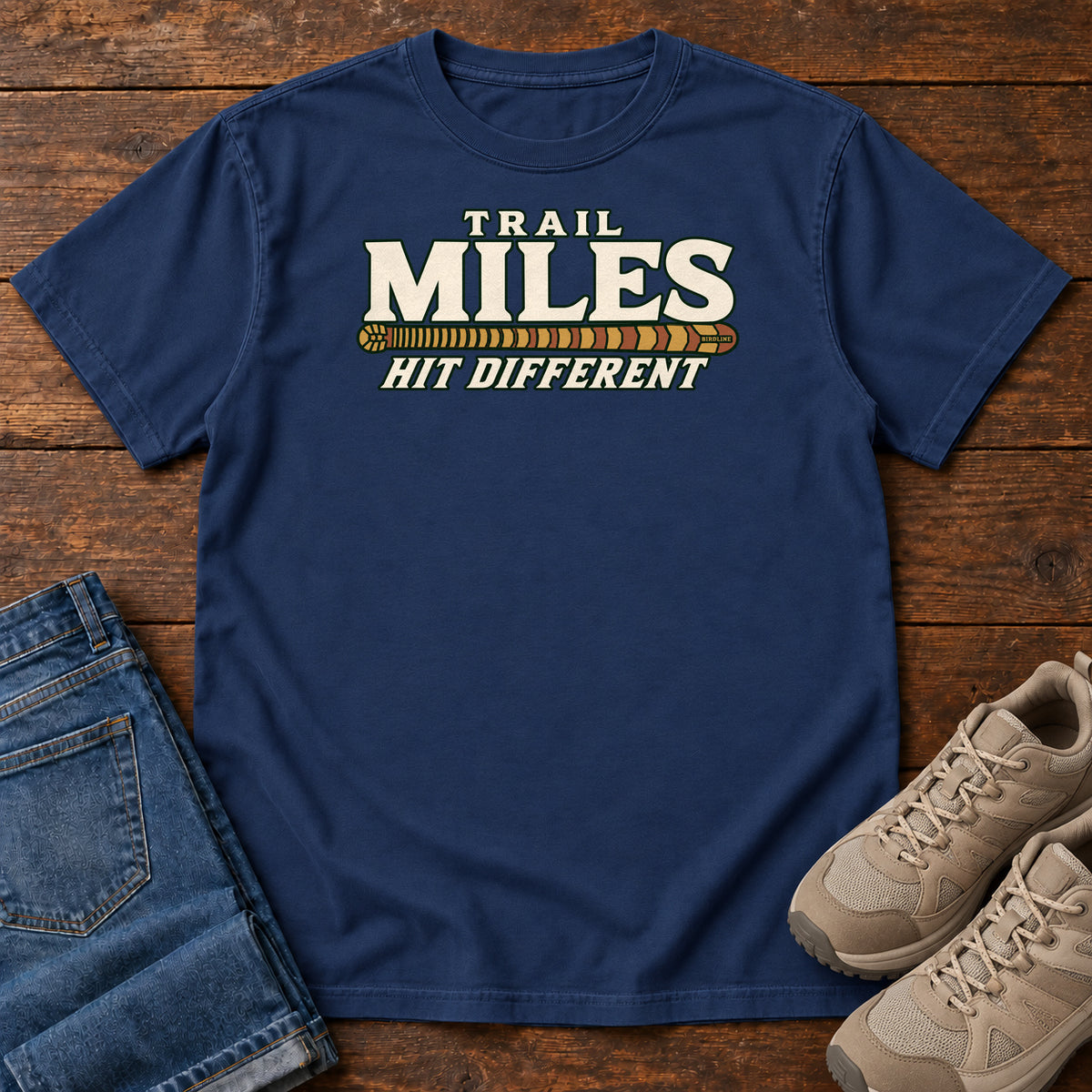True Navy Trail Miles Hit Different T-Shirt with cream lettering and a gold-to-rust patterned stripe, front flat lay on wood.