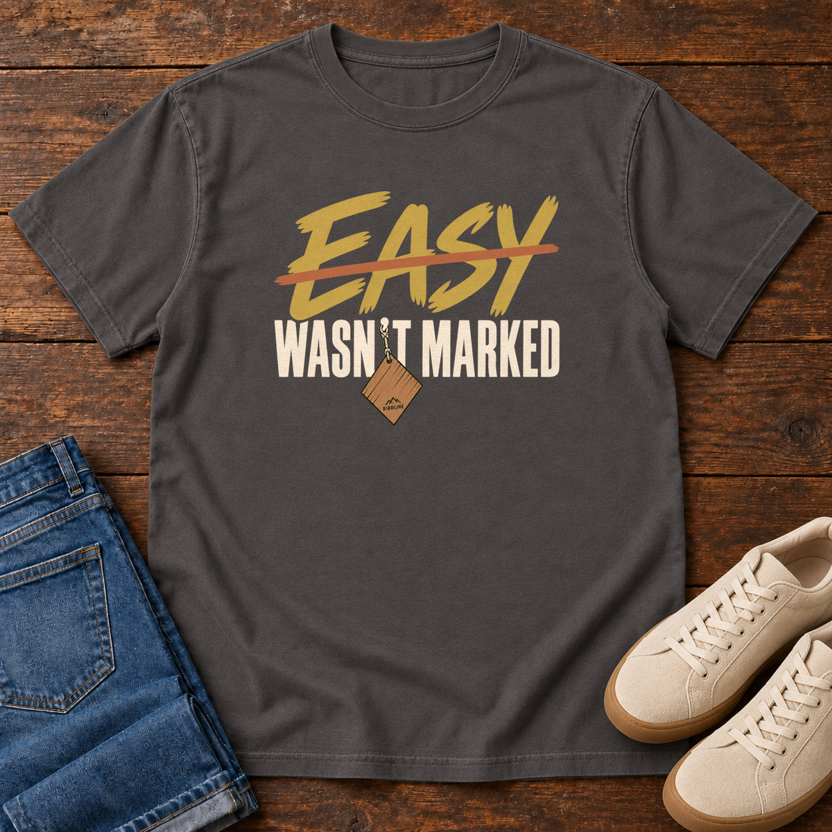 Graphite Easy Wasn't Marked T-Shirt with gold Easy lettering crossed out in orange above cream Wasn't Marked text, front flat lay on wood.