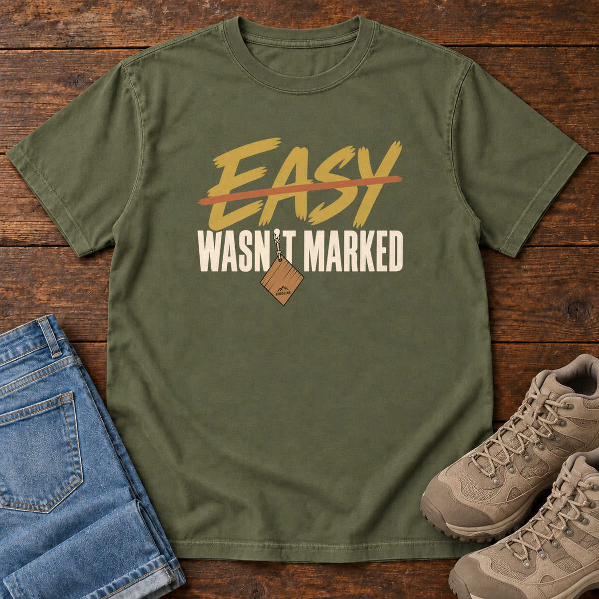 Hemp Easy Wasn't Marked T-Shirt with gold Easy lettering crossed out in orange above cream Wasn't Marked text, front flat lay on wood.