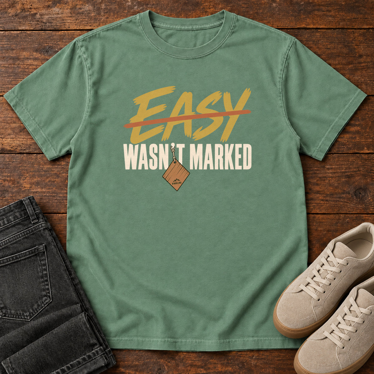 Light Green Easy Wasn't Marked T-Shirt with gold Easy lettering crossed out in orange above cream Wasn't Marked text, front flat lay on wood.